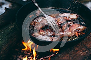 Slices of fried bacon in a pan. Food in a forest camp. Cooking on fire. Picnic in the nature. Grilled food on nature. Fork taking