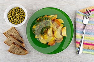 Slices of fried potatoes in plate with dill, bowl with green peas, bread, fork on napkin on table. Top view