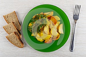 Slices of fried potatoes in plate with dill, bread, fork on table. Top view