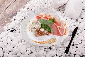 Slices of ham, walnuts and cheese on a plate. A knife and fork near a dish on a white stones background. Luxury dinner.