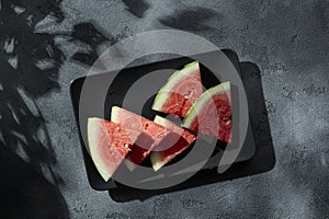 Slices of juicy watermelon on the black plate on the concrete background with summer sun shadows. Copy space. Top view