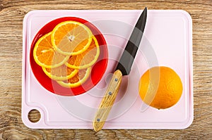 Slices of oranges in red saucer, knife, full orange on cutting board on wooden table. Top view