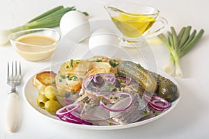 Slices of pickled herring with red onion, fermented cucumber, fried potatoes, olives, mustard sauce, boiled eggs and herbs