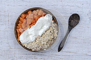Slices of sweet papaya with oat flakes and white yogurt in coconut bowl on white wooden background, close up