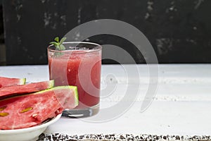 Slices of ripe watermelon on a plate and red watermelon juice in a glass on a wooden white background, and a black wall - side