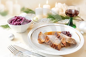 Slices of roast pork with crust on a plate served on a festive table with red cabbage, red wine, white candles and Christmas