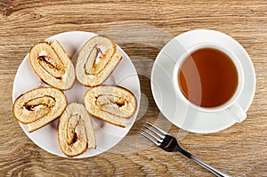 Slices of roll with cream and jam in white plate, fork, tea in cup on saucer on table. Top view