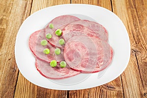 Slices of sliced ham and green peas on a white plate on a wooden table