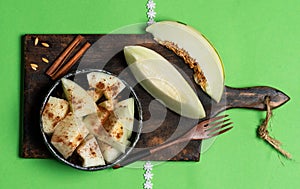 Slices and small pieces of ivory gaya melon sprinkled with cinnamon in a bowl, on a cutting board and a green background. Top view