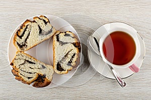 Slices of sweet bun with poppy in plate, cup of tea, spoon on saucer on wooden table.
