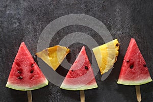 Slices of water-melon and pineapple on popsickle stick on gray background flat lay