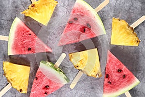 Slices of water-melon and pineapple on popsickle stick on gray background flat lay