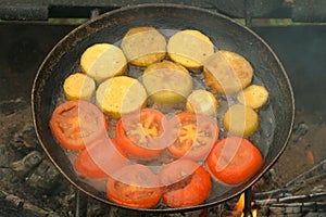 Slices of zucchini and tomatoes are frying on frying pan with oil in chargrill.