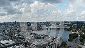 Sliding aerial footage of water surface surrounded by buildings and with fountain in middle. Fly along Binnenalster lake