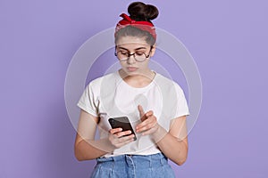 Slightly hunched woman with hair bun using mobile phone with unhappy facial expression, holds smart phone, texting in social