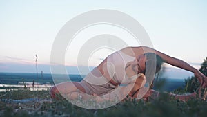 Slim adult woman doing yoga on the hill at early evening - stretching to the side touching her toes