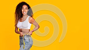 Slim african american woman measuring waist with tape, yellow backdrop