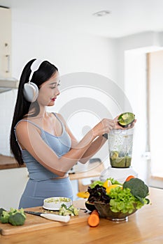 A slim Asian woman in gym clothes and headphones is making her healthy green smoothie