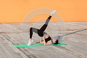 Slim athletic girl in tight sportswear, black pants and top, practicing yoga, doing one legged bridge pose with leg raise