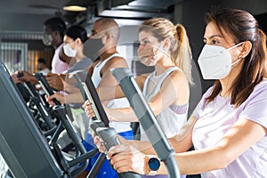 Slim athletic people in protective masks running on treadmill in fitness club