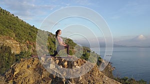 Slim blond girl stands in warrior pose on stone against hill sea