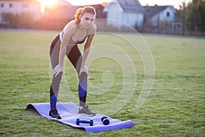 Slim blonde girl plays sports and performs yoga poses in summer grass covered stadium on a sunset background. Woman doing