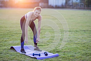 Slim blonde girl plays sports and performs yoga poses in summer grass covered stadium on a sunset background. Woman doing