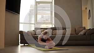 Slim, blonde woman sits on mat, does exercises and stretches by leaning torso forward at morning in bright living room