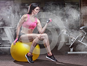 Slim, bodybuilder girl, lifts heavy dumbbell sits on a yoga ball in the gym. Sport concept, fat burning and a healthy lifestyle