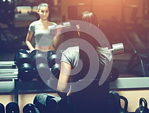 Slim, bodybuilder girl, lifts heavy dumbbell standing in front of the mirror while training in the gym.