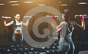 Slim, bodybuilder girl, lifts heavy dumbbell standing in front of the mirror while training in the gym.