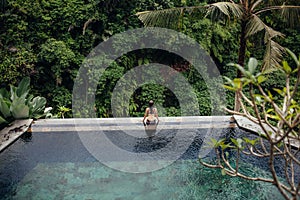 Slim brunette woman in swimsuit relaxing on edge infinity pool in jungle. Palms around and crystal clean water