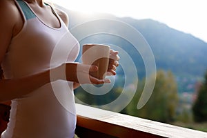 Slim caucasian woman holds cup of tea in her hands at mountain resort. Sports girl with hot coffee mug at wooden balcony