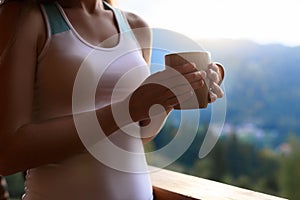 Slim caucasian woman holds cup of tea in her hands at mountain resort. Sports girl with hot coffee mug at wooden balcony