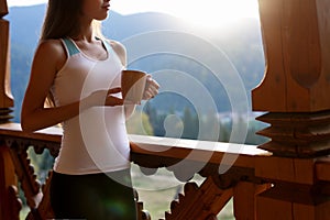 Slim caucasian woman holds cup of tea in her hands at mountain resort. Sports girl with hot coffee mug at wooden balcony