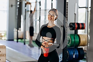 Slim dark-haired girl dressed in black sportswear stands with water in her hand near the sport equipment in the gym