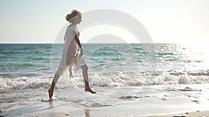 Slim excited beautiful woman with tattoo on leg running in sunlight on sandy Mediterranean sea beach. Wide shot side