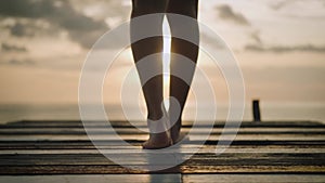 Slim female legs and feet back view of woman silhouette walking barefoot on wooden jetty pier and watching sunset.