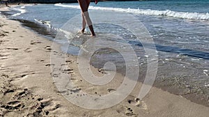 Slim female legs and feet walking along sea water waves on sandy beach. Pretty woman walks at seaside surf.