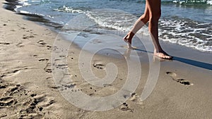 Slim female legs and feet walking along sea water waves on sandy beach. Pretty woman walks at seaside surf.