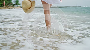 Slim female legs and feet walking along sea water waves on sandy beach. Pretty woman walks at seaside surf wearing white