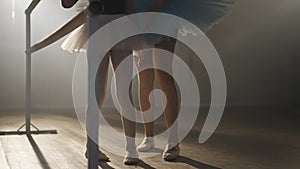 Slim female legs of young woman and little girl in tutu at ballet barre in backlit fog. Unrecognizable teacher and