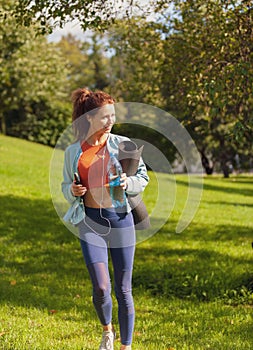slim fit caucasian woman in colorful fitness outfit walking in the park with yoga mat bottle of watter and smartphone