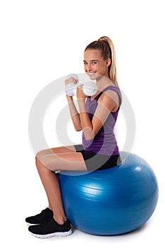 Slim and fit teen girl sitting a swiss blue ball and holding white dumbbells . Full length shot on white background