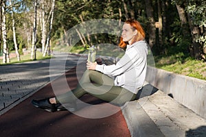 slim ginger woman runner in sport clothes sitting in park, drinking water. Healthy fitness lifestyle