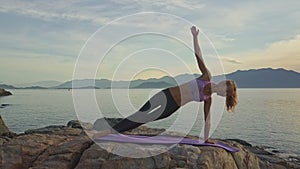 Slim girl balances on left arm in yoga pose on rock