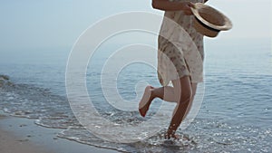 Slim girl running ocean water holding hat summer day. Closeup woman legs walking
