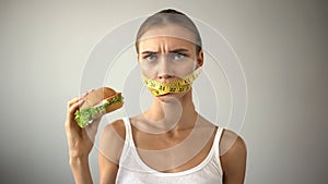 Slim girl with taped mouth fights with temptation to eat burger, dieting