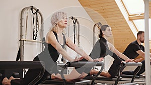 Slim good looking senior woman doing core exercises while taking pilates lesson on a reformer machine, by female