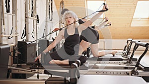 Slim good looking senior woman doing core exercises while taking pilates lesson on a reformer machine, by female